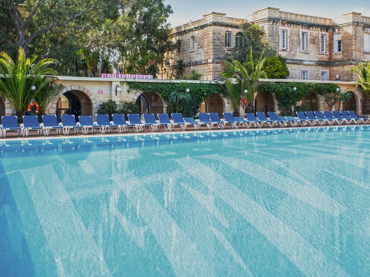 Large outdoor swimming pool surrounded by sun loungers and Mediterranean-style buildings at Sprachcaffe Malta.